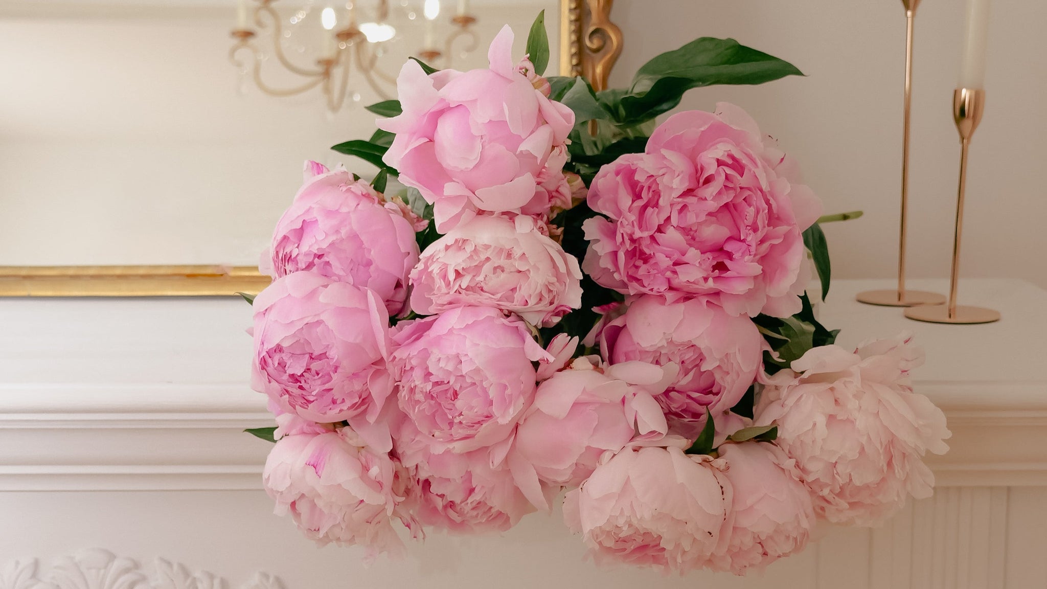 Pink peony flowers on a decorative fireplace mantel with candles and a chandelier in the background.